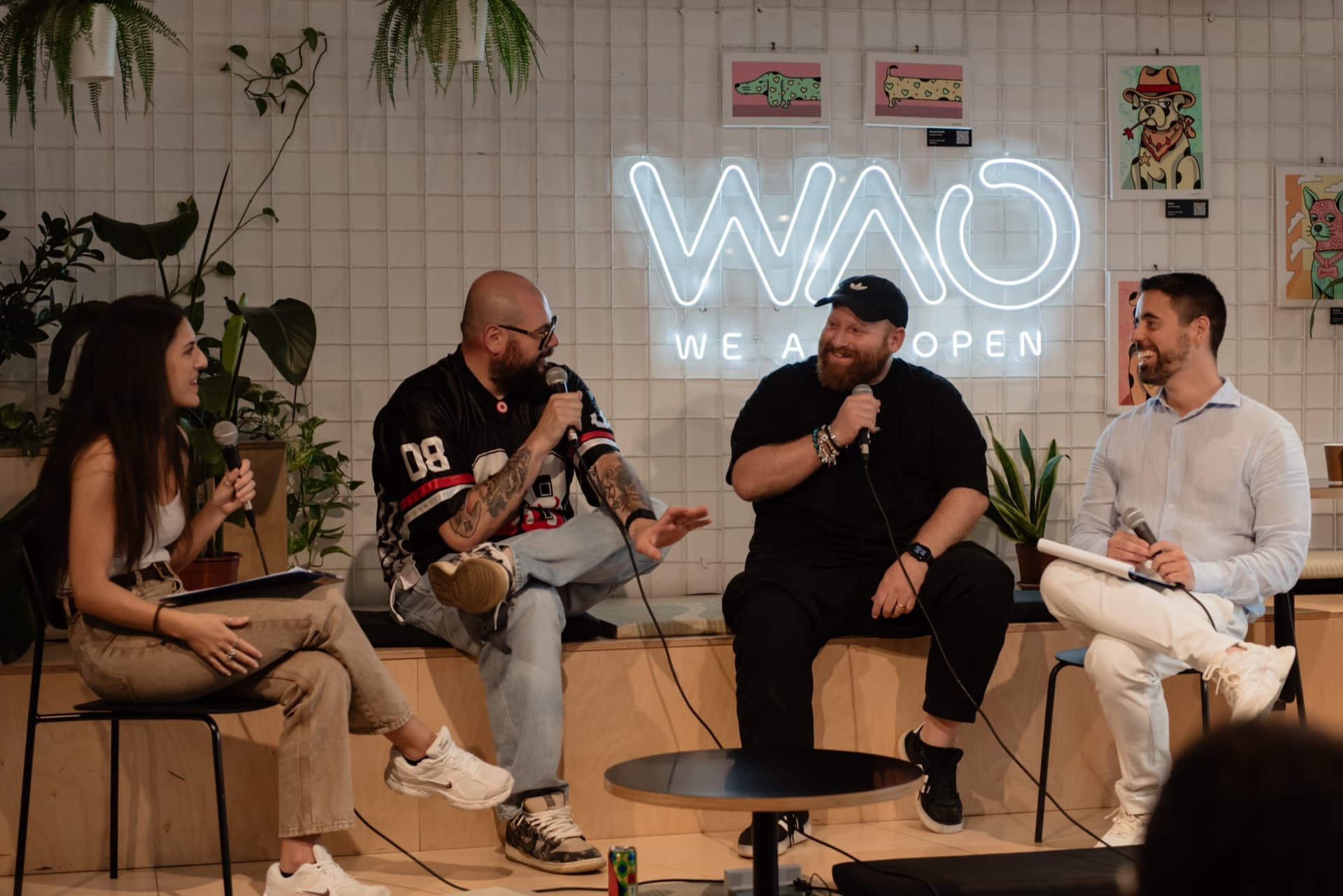 Four speakers with microphones at a talk, under the WAO neon sign