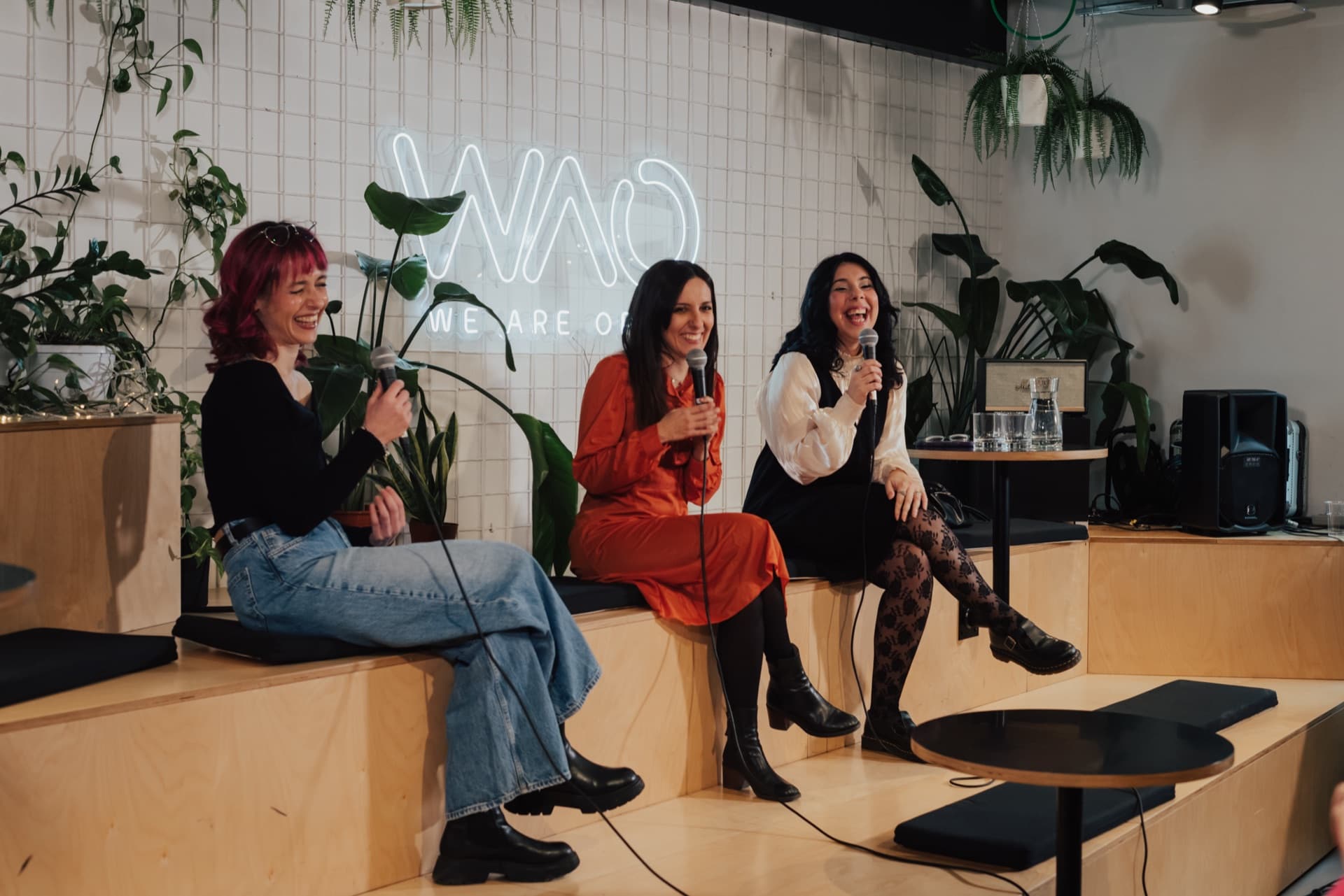 Three speakers holding microphones during a talk on the bleachers, under the WAO neon sign